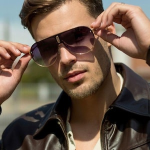 May include: Sunglasses with a dark gradient lens and gold-colored frames. The sunglasses are held up by a person wearing a brown leather jacket and a light-colored shirt. The background is a blurred blue sky.