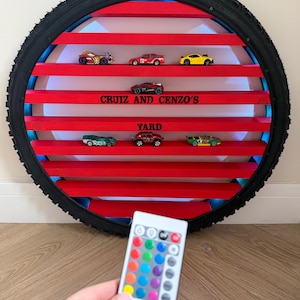May include: A circular display shelf made from a tyre rim, with red shelves holding toy cars. The text "CRUIZ AND CENZO'S YARD" is visible. A remote control with colour options is in the foreground.