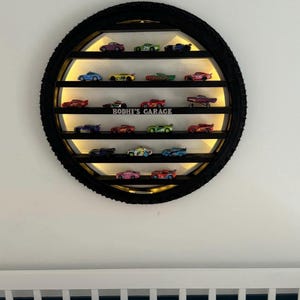 May include: A round, black shelf unit crafted from a tyre, showcasing a collection of toy cars. The shelf features several levels with integrated lighting. The text "BODHI'S GARAGE" is displayed. The toy cars are in various colours and designs.
