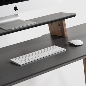 May include: A modern black desk with wooden legs and a matching monitor stand. A white keyboard, mouse, and a mug are on the desk. The desk is designed for a home office or workspace.