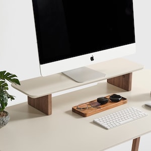Puede incluir: Un escritorio moderno con una superficie color crema y patas de madera. Un monitor de ordenador se encuentra sobre un soporte a juego. Los accesorios incluyen un teclado, un rat&oacute;n, una taza y una peque&ntilde;a planta en maceta. Una bandeja de madera contiene gafas y peque&ntilde;os recipientes.