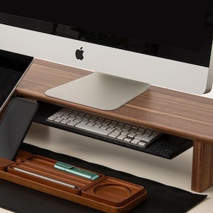 K&ouml;nnte beinhalten: Ein h&ouml;lzerner Schreibtisch-Organizer mit einer schwarzen Leder-Schreibtischunterlage, einem schwarzen Mauspad, einem schwarzen Computermonitor, einer schwarzen Tastatur, einem schwarzen Tablet, einem schwarzen Smartphone, einem schwarzen Headset und einem schwarzen Laptop-St&auml;nder.