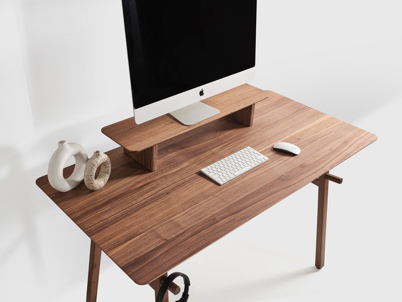 May include: A rectangular wooden desk with a natural grain finish, featuring a matching elevated monitor stand. On the desk surface are a white computer keyboard, a white computer mouse, two white ceramic vases, and a black computer monitor.