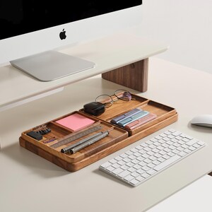 Puede incluir: Una moderna configuraci&oacute;n de escritorio con un monitor de computadora, teclado y rat&oacute;n blancos. Una bandeja organizadora de madera contiene &uacute;tiles de oficina como bol&iacute;grafos, un bloc de notas y gafas. El escritorio es de color claro con un soporte de monitor de madera.