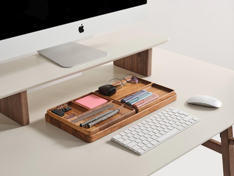 Puede incluir: Una moderna configuraci&oacute;n de escritorio con un monitor de computadora, teclado y rat&oacute;n blancos. Una bandeja organizadora de madera contiene &uacute;tiles de oficina como bol&iacute;grafos, un bloc de notas y gafas. El escritorio es de color claro con un soporte de monitor de madera.