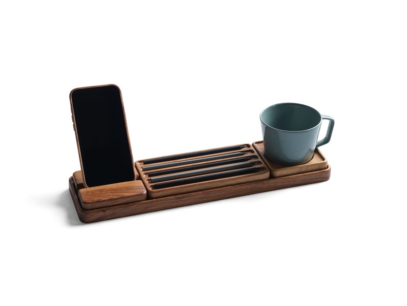 May include: A rectangular wooden desk organizer featuring a slot for a smartphone, a recessed tray holding four black pencils, and a square coaster section supporting a solid light blue ceramic mug with a handle, all crafted from dark wood.
