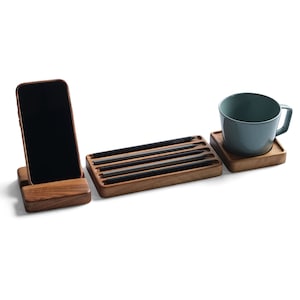 May include: A wooden desk organizer set consisting of a smartphone stand holding a black phone, a rectangular tray containing four black pencils, and a square wooden coaster supporting a light blue ceramic mug with a handle.