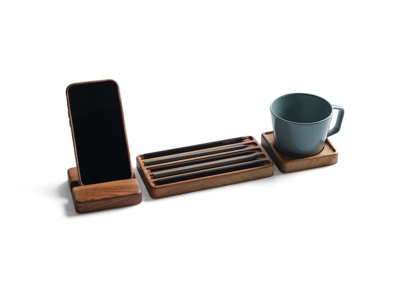 May include: A wooden desk organizer set consisting of a smartphone stand holding a black phone, a rectangular tray containing four black pencils, and a square wooden coaster supporting a light blue ceramic mug with a handle.