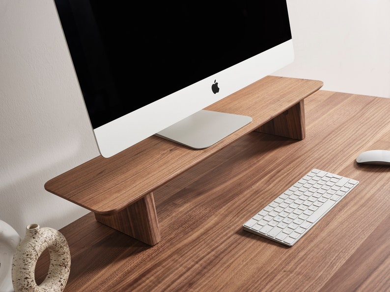May include: A rectangular wooden monitor stand with a smooth, dark brown wood grain finish and two solid side supports. The stand is positioned on a matching wooden desk surface, holding a silver computer monitor with a black screen.