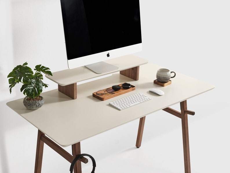 Puede incluir: Un escritorio moderno con una superficie color crema y patas de madera. Un monitor de ordenador se encuentra sobre un soporte a juego. Los accesorios incluyen un teclado, un rat&oacute;n, una taza y una peque&ntilde;a planta en maceta. Una bandeja de madera contiene gafas y peque&ntilde;os recipientes.