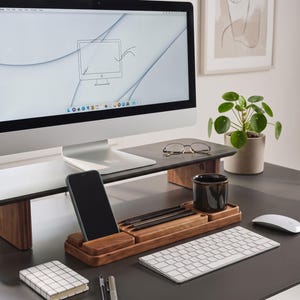 May include: A wooden monitor stand and a matching wooden desk organizer with a smartphone slot, pen tray, and cup holder. The setup includes a computer monitor, a white keyboard, a white mouse, a grid-patterned notebook, two pens, and a pair of glasses.