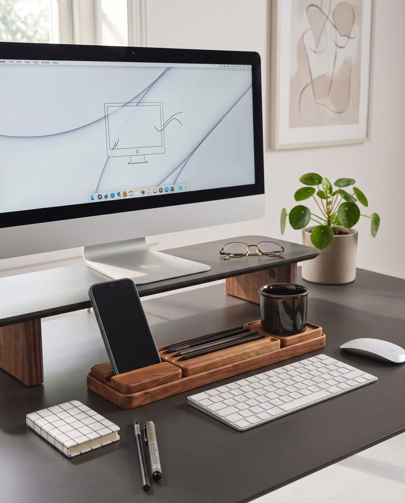 May include: A wooden monitor stand and a matching wooden desk organizer with a smartphone slot, pen tray, and cup holder. The setup includes a computer monitor, a white keyboard, a white mouse, a grid-patterned notebook, two pens, and a pair of glasses.