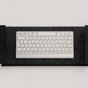 K&ouml;nnte beinhalten: Eine wei&szlig;e Tastatur mit schwarzen Tasten liegt auf einem schwarzen, mit Filz ausgekleideten Regal. Das Regal ist an einer wei&szlig;en Wand befestigt.