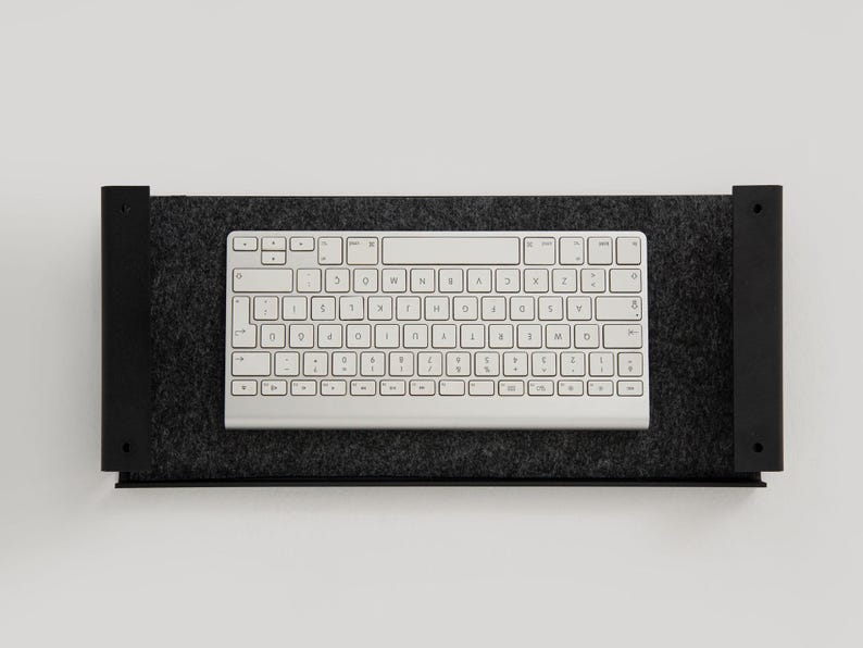 K&ouml;nnte beinhalten: Eine wei&szlig;e Tastatur mit schwarzen Tasten liegt auf einem schwarzen, mit Filz ausgekleideten Regal. Das Regal ist an einer wei&szlig;en Wand befestigt.