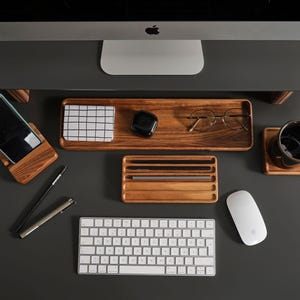 May include: A wooden desk organizer set including a rectangular tray, a phone stand, a pen holder with four slots, and a square coaster. The set is arranged with a smartphone, a grid-patterned notepad, black earbuds, glasses, and two pens.
