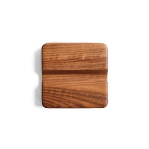 May include: A square wooden phone stand featuring a rich brown grain pattern and rounded corners. The stand has a horizontal groove cut across the center, designed to hold a mobile device in a stable, upright position for viewing.