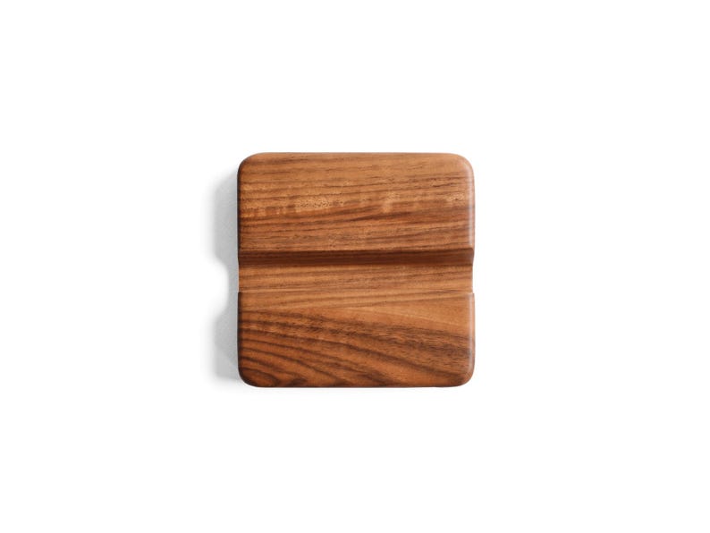 May include: A square wooden phone stand featuring a rich brown grain pattern and rounded corners. The stand has a horizontal groove cut across the center, designed to hold a mobile device in a stable, upright position for viewing.