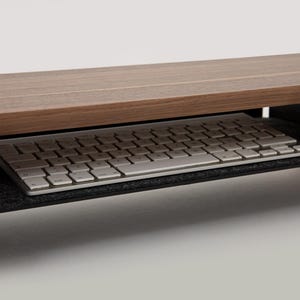 K&ouml;nnte beinhalten: Ein Monitorst&auml;nder aus Holz mit einer ausziehbaren Tastaturablage. Der St&auml;nder ist braun und die Ablage ist schwarz mit einem grauen Filzbezug. Eine wei&szlig;e Tastatur liegt in der Ablage.