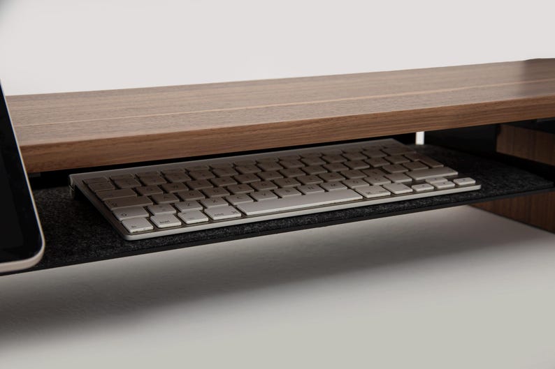 K&ouml;nnte beinhalten: Ein Monitorst&auml;nder aus Holz mit einer ausziehbaren Tastaturablage. Der St&auml;nder ist braun und die Ablage ist schwarz mit einem grauen Filzbezug. Eine wei&szlig;e Tastatur liegt in der Ablage.