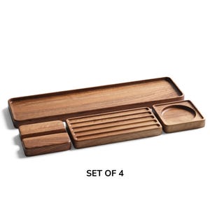 May include: A set of four brown wooden trays featuring a long rectangular tray, a small rectangular tray with four parallel grooves, a square tray with a circular indentation, and a small rectangular block. The text SET OF 4 is visible.