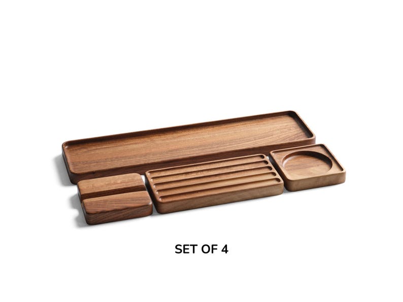 May include: A set of four brown wooden trays featuring a long rectangular tray, a small rectangular tray with four parallel grooves, a square tray with a circular indentation, and a small rectangular block. The text SET OF 4 is visible.