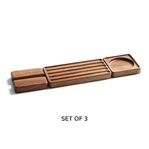 May include: A set of three brown wooden trays with a natural grain finish. The set includes two rectangular blocks, one long rectangular tray with five parallel grooves, and one square tray featuring a circular indentation. The text SET OF 3 is visible.