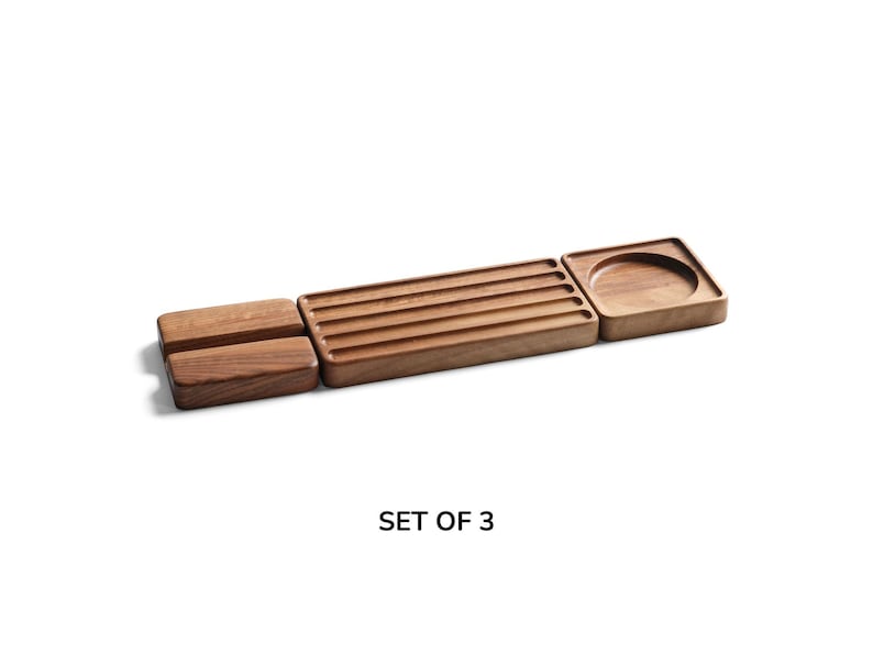 May include: A set of three brown wooden trays with a natural grain finish. The set includes two rectangular blocks, one long rectangular tray with five parallel grooves, and one square tray featuring a circular indentation. The text SET OF 3 is visible.