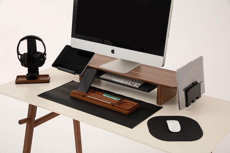 K&ouml;nnte beinhalten: Ein h&ouml;lzerner Schreibtisch-Organizer mit einer schwarzen Leder-Schreibtischunterlage, einem schwarzen Mauspad, einem schwarzen Computermonitor, einer schwarzen Tastatur, einem schwarzen Tablet, einem schwarzen Smartphone, einem schwarzen Headset und einem schwarzen Laptop-St&auml;nder.