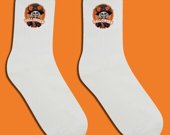 Puede incluir: Par de calcetines blancos con diseño de Halloween. Cada calcetín muestra un gráfico circular con un esqueleto con sombrero, rodeado de flores naranjas y la palabra "HALLOWEEN".