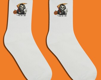 Puede incluir: Par de calcetines blancos con un dibujo animado de la Parca con una guadaña y una calabaza. La Parca hace el signo de la paz. El texto "I LOVE MY JOB" está impreso debajo.