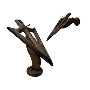 May include: A wooden arrowhead wall mount with a dark brown stain. The arrowhead is mounted on a curved wooden base with a circular base. The arrowhead is angled upwards and the base is angled downwards. The arrowhead is made of wood and has a realistic arrowhead shape.