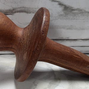 Pode incluir: Uma ferramenta de massagem de madeira marrom. Possui uma cabeça arredondada, uma seção intermediária alargada e uma extremidade pontiaguda. A ferramenta é feita de uma única peça de madeira. O design sugere uso terapêutico.
