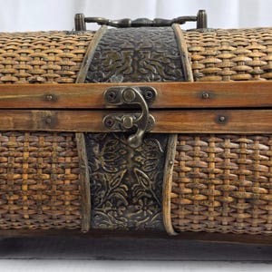 May include: A rectangular, woven wicker chest with a hinged lid. The chest features a dark brown wooden frame and decorative metal accents. A metal clasp secures the lid, and a handle is on top.
