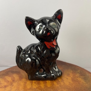 May include: A glossy black ceramic cat figurine with red accents on the ears and a red bow tie. The cat is in a sitting position, with its tail curled to the side. The figurine is on a brown surface.