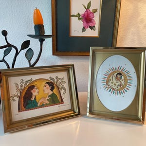 May include: Three framed artworks with gold frames are displayed on a white surface. One features a watercolor of a pink flower, another depicts two figures, and the third shows a person surrounded by a sunburst design. A black candle holder is on the left.