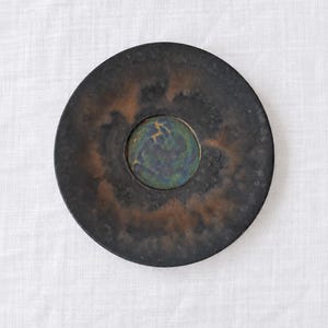 May include: A circular ceramic plate with a dark, matte black and rusted brown exterior finish. The center features a recessed, circular inlay with a glossy, mottled blue and green glaze, containing a small, faint gold-colored deer silhouette.