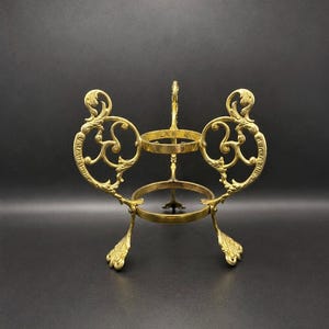May include: A gold-toned metal stand featuring two circular rings stacked vertically, supported by three ornate, curved legs with leaf-like feet and decorative scrollwork side panels that connect the upper and lower ring sections.