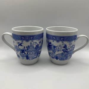 Op de afbeelding: Twee witte keramische mokken met blauwe Willow-patroonontwerpen. De mokken hebben een afgeronde vorm met een handvat en een blauwe decoratieve rand. Het blauwe patroon toont een landschap met gebouwen, bomen en figuren.