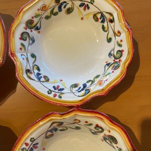 May include: A set of off-white ceramic bowls with a scalloped edge. The rims are decorated with a yellow band, a red border, and a painted floral design in green, blue, and red.