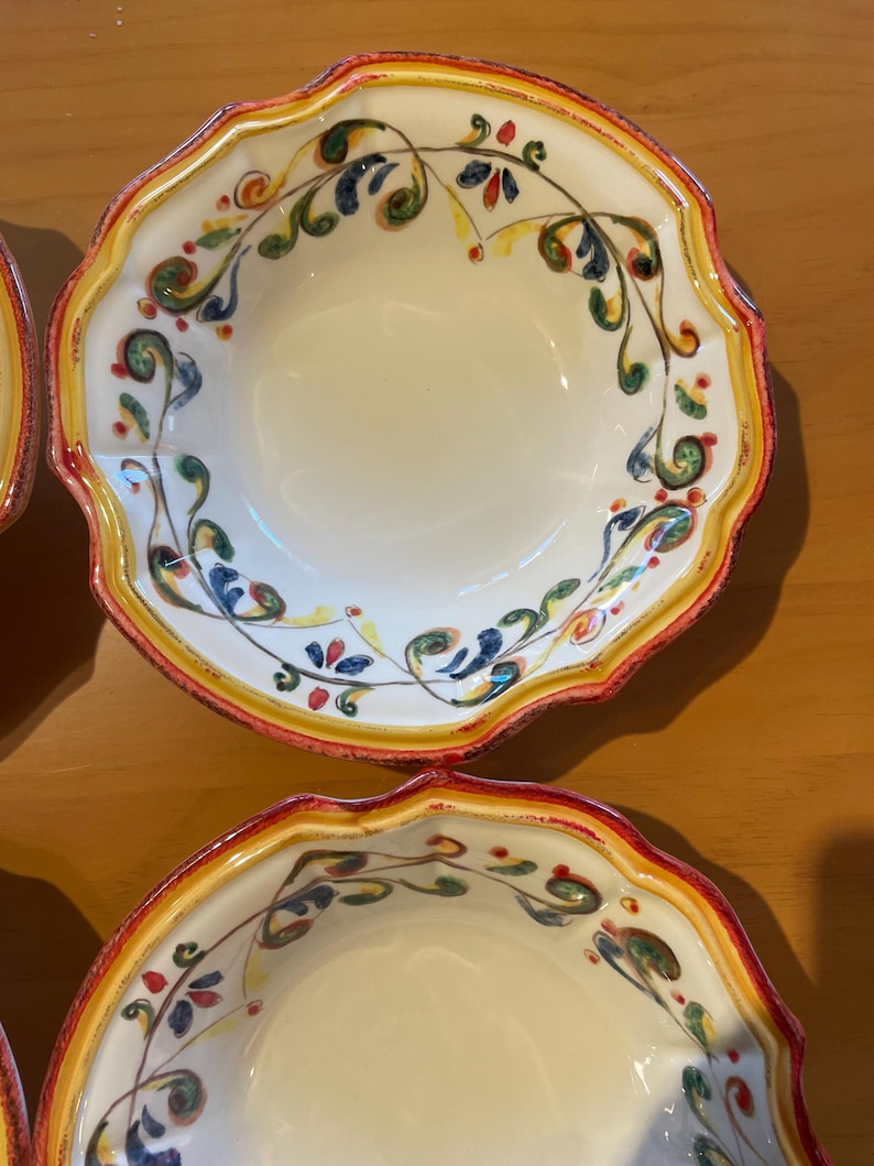May include: A set of off-white ceramic bowls with a scalloped edge. The rims are decorated with a yellow band, a red border, and a painted floral design in green, blue, and red.