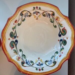 May include: A white ceramic serving bowl with a scalloped edge. The rim is decorated with a red and yellow border. The bowl features a hand-painted floral design in green, blue, and red. The bowl is likely for serving food.