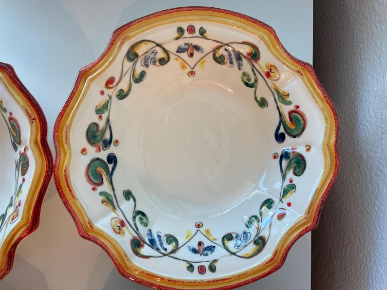 May include: A white ceramic serving bowl with a scalloped edge. The rim is decorated with a red and yellow border. The bowl features a hand-painted floral design in green, blue, and red. The bowl is likely for serving food.
