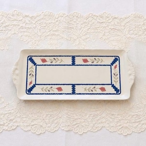 May include: A rectangular ceramic serving tray with a cream-colored base and scalloped handles. The tray features a central rectangular border in dark blue, accented with small red floral motifs and green leaves along the edges of the frame.