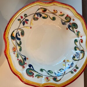 May include: A white ceramic serving bowl with a scalloped edge, featuring a red, yellow, and green floral design. The bowl has a wide rim with a red border and a yellow inner edge. The floral pattern includes blue, green, and red accents.