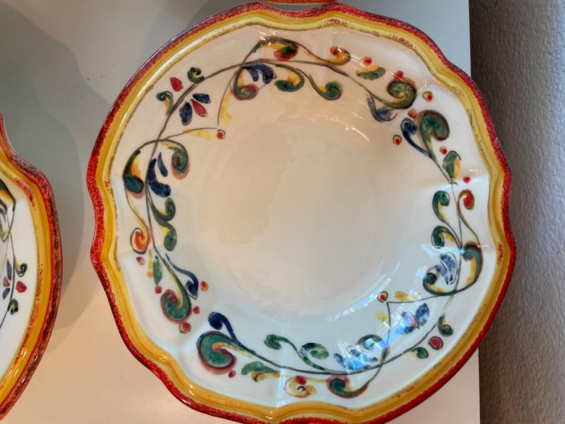 May include: A white ceramic serving bowl with a scalloped edge, featuring a red, yellow, and green floral design. The bowl has a wide rim with a red border and a yellow inner edge. The floral pattern includes blue, green, and red accents.
