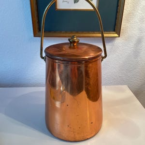 May include: A copper bucket with a lid and a brass handle. The bucket is cylindrical and has a small brass knob on the lid. The handle arches over the top. The bucket is on a white surface. The bucket is likely vintage.