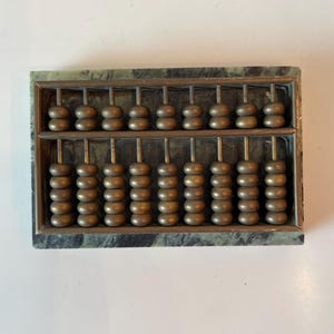 May include: A vintage abacus with a green marble frame and brass-coloured beads. The abacus has two rows of beads on top and five rows of beads on the bottom. The beads are arranged on metal rods.