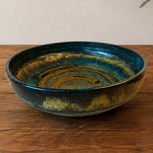 May include: A round ceramic bowl featuring a deep teal and dark blue glaze with swirling golden-brown and ochre accents. The interior surface displays a smooth, glossy finish with concentric color patterns, while the exterior has a dark, mottled texture.