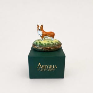 以下が含まれることがあります： 手描きの茶色と白のコーギー犬のフィギュアが緑の草の土台に乗ったリモージュ磁器の小物入れ。この箱は、前面に金色の文字でArtoria Limogesと印刷された濃い緑色の正方形の箱の上に置かれています。