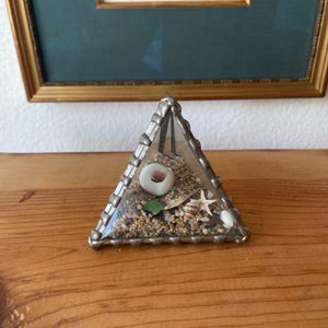 May include: A triangular glass terrarium with a silver-colored metal frame. The terrarium contains sand, seashells, and a small green piece of sea glass. A framed floral print hangs in the background.